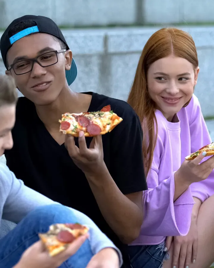 Teen friends sitting on steps outside, eating pizza and laughing