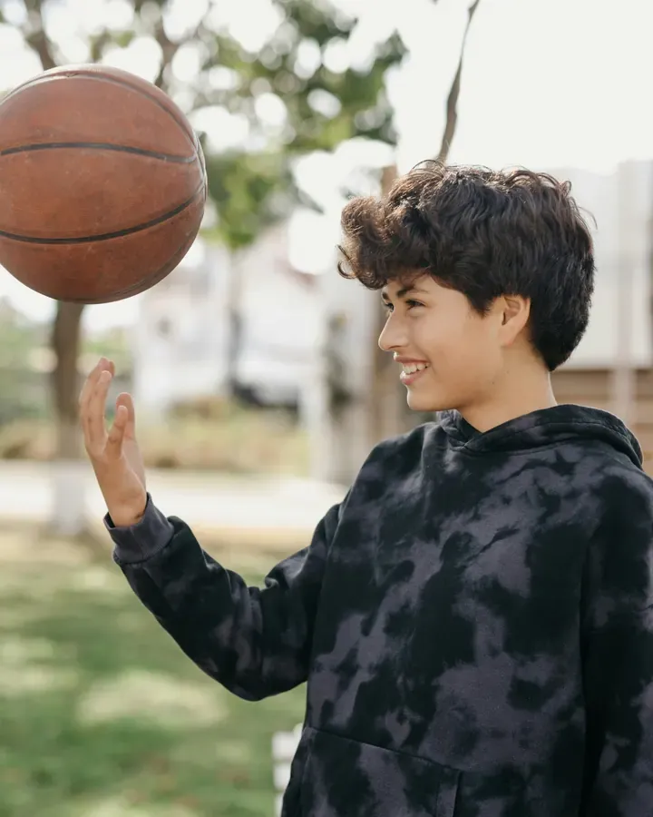 A smiling teen in a hoodie spinning a basketball on one finger outdoors